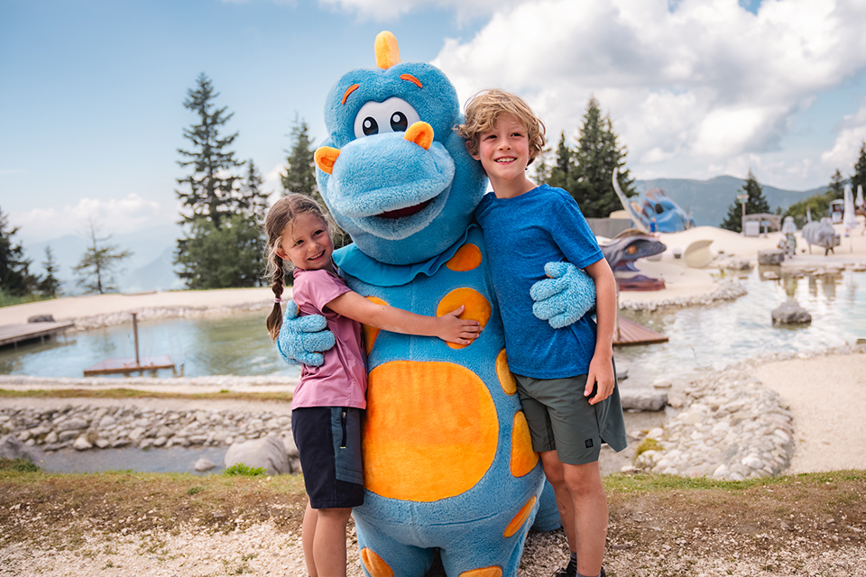 Bergbahn Steinplatte by Klaus Listl Zwei Kinder mit dem Maskottchen der Steinplatte und im Hintergrund die Bergkulisse, Wasser und Dions