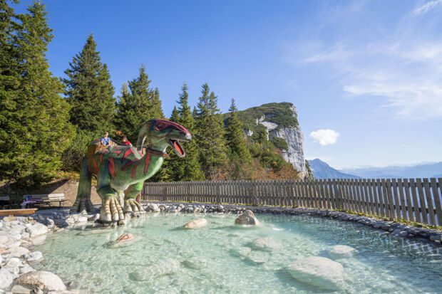 TriassicPark_Steinplatte_Waidring_©defrancesco (4) (1) Kinder spielen auf Dinosaurier-Figur im Wasser im Triassic Park Steinplatte mit Bergblick