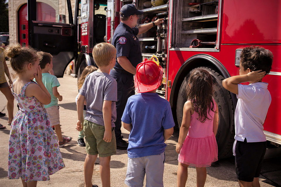 Mehrere Kinder stehen an einem roten Feuerwehrauto und schauen einem Feuerwehrmann zu, der ihnen die Ausrüstung in den geöffneten Seitenfächern zeigt.